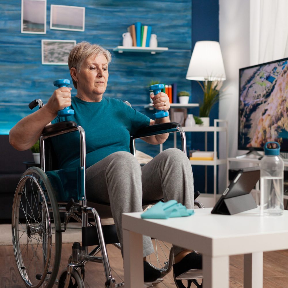Handicapped senior woman holding trainer dumbbells doing arms exercise working at body persistence training in living room. Disabled pensioner looking at online muscles workout course on tablet